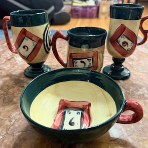 May include: A set of three ceramic mugs and one wide bowl featuring a cream-colored base with dark green rims and handles. Each piece is decorated with a hand-painted red square frame containing a small blue and black abstract symbol.