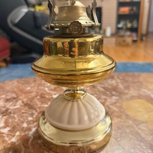 Puede incluir: Una pequeña lámpara de aceite antigua con acabado de latón. La lámpara presenta una base blanca con diseño acanalado y una parte superior de latón con mecha. La lámpara está diseñada para colocarse sobre una mesa u otra superficie plana.