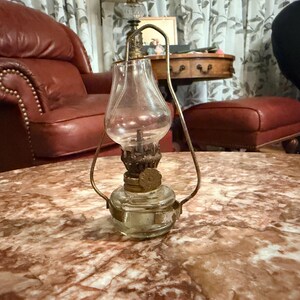May include: A small, clear glass oil lamp with a brass handle and accents. The lamp has a rounded base and a bell-shaped glass chimney. The lamp is sitting on a marble-patterned table. The lamp is an antique.