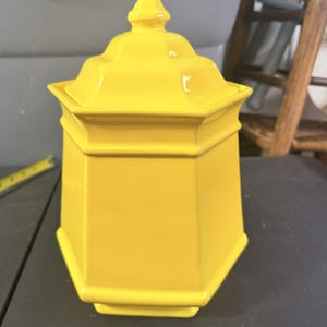 May include: A bright yellow ceramic canister with a hexagonal body and a matching removable lid featuring a pointed finial top. The container has a smooth, glossy finish and a structured, geometric shape with defined edges and a flat base.