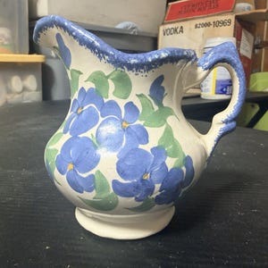 May include: A cream-colored ceramic pitcher featuring a hand-painted pattern of blue flowers with yellow centers and green leaves. The rim and the handle are decorated with a textured, sponged blue paint finish.
