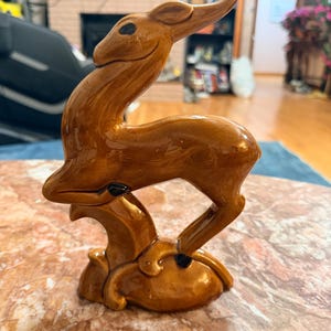 May include: A brown ceramic deer figurine with a glossy finish. The deer is posed in a leaping position, with its head and neck arched gracefully. The figurine is mounted on a decorative base.