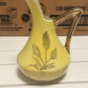 May include: A small yellow ceramic creamer pitcher featuring a gold metallic handle and rim. The body of the pitcher is decorated with a gold wheat stalk illustration and a textured gold band around the base, with a curved spout for pouring.