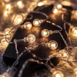 May include: A string of warm white fairy lights with clear, round bulbs. The bulbs have a textured, frosted finish and are strung on a clear wire.