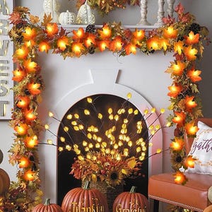 May include: A decorated fireplace with a garland of orange maple leaf lights. The garland is draped with autumn foliage. Two pumpkins with the words "Thankful" and "Grateful" are in front of the fireplace, creating a seasonal display.