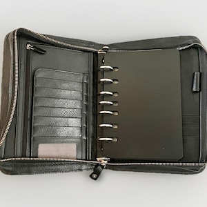 May include: Open black leather organiser with a silver zip. The interior features a ring binder, card slots, and a pen holder. The organiser is open, revealing a black notepad and additional storage compartments.