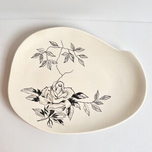 May include: Cream-colored, oval-shaped serving platter with a textured surface. The platter features a black and white floral design of a rose and leaves. The design is centered on the platter.