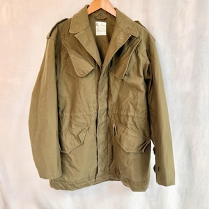 Puede incluir: Chaqueta verde oliva de estilo militar con cuello, cuatro bolsillos delanteros y puños abotonados. La chaqueta está colgada de una percha de madera. Una etiqueta es visible cerca del cuello. La chaqueta está hecha de una tela resistente.