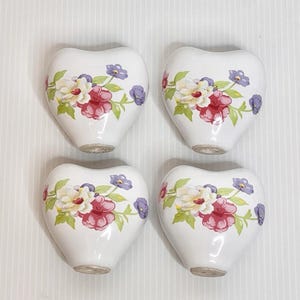 Può includere: Quattro pomelli in ceramica bianchi a forma di cuore con disegni floreali. Ogni pomello presenta un gruppo di fiori rossi, bianchi e viola con foglie verdi. I pomelli sono disposti a quadrato.