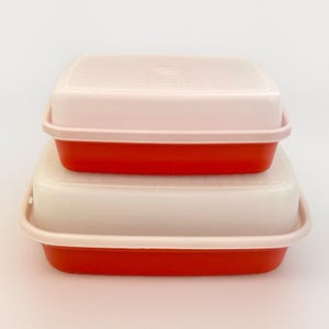 Puede incluir: Dos recipientes cuadrados para guardar alimentos apilados. Los recipientes tienen bases rojas y tapas de color rosa claro. Las tapas tienen una superficie texturizada. Los recipientes son de plástico.