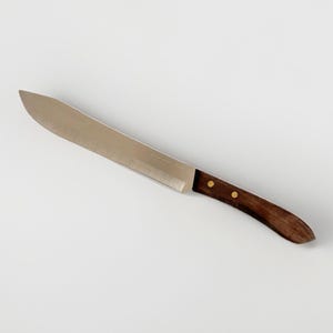 Peut inclure: Une grande machette en acier inoxydable avec un manche incurvé en bois brun foncé. La lame est longue et large, conçue pour hacher. Deux rivets en laiton fixent le manche à la lame.