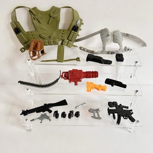 Puede incluir: Una colección de accesorios militares de juguete, que incluye un chaleco verde, una pequeña mochila marrón y varias armas de juguete negras y grises. Otros artículos incluyen un cañón rojo, un arma naranja y rodilleras grises. Los artículos se exhiben en estantes de acrílico transparente.