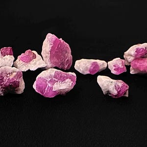 May include: A collection of raw gemstones, showcasing a vivid pink colour within a matrix of off-white rock. The stones are arranged against a black backdrop. These uncut gems offer a natural aesthetic.