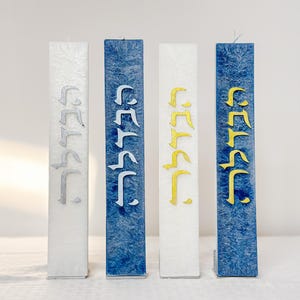 May include: Four rectangular candles in a row. The candles are white, blue, and white with gold lettering. The candles have a textured appearance and feature stylized lettering in silver and gold.