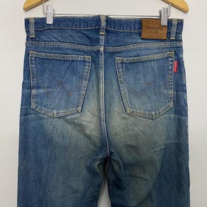 Może przedstawiać: Zbliżenie na tył niebieskich dżins&oacute;w Wrangler. Dżinsy mają dwie tylne kieszenie z wszytym logo Wrangler. Na pasku widoczna jest brązowa sk&oacute;rzana naszywka z logo Wrangler.