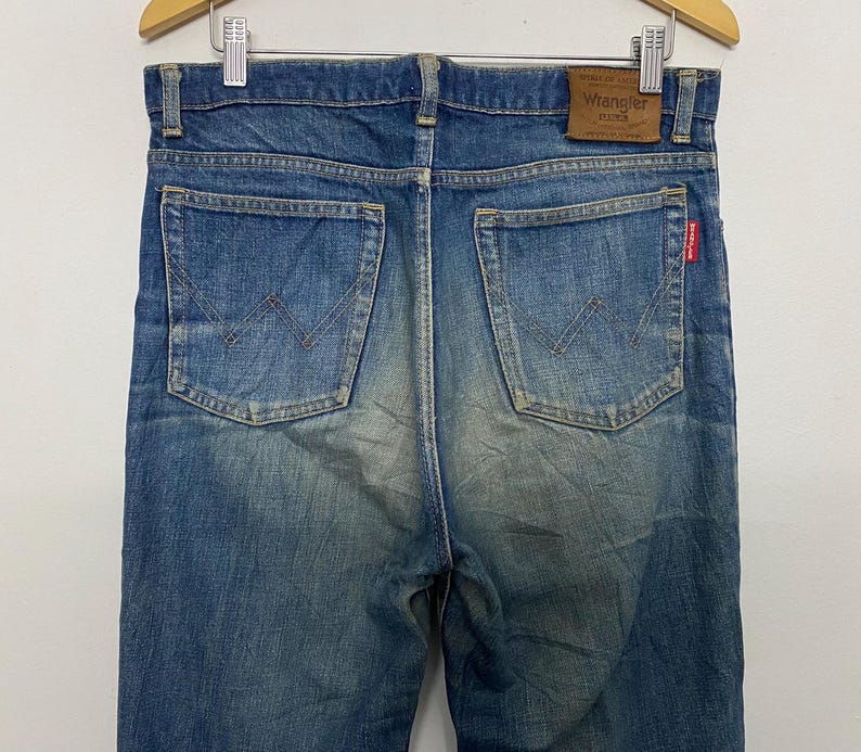 Może przedstawiać: Zbliżenie na tył niebieskich dżins&oacute;w Wrangler. Dżinsy mają dwie tylne kieszenie z wszytym logo Wrangler. Na pasku widoczna jest brązowa sk&oacute;rzana naszywka z logo Wrangler.