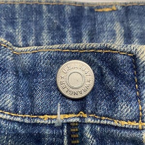 Może przedstawiać: Zbliżenie niebieskich dżins&oacute;w z srebrnym guzikiem i zamkiem błyskawicznym. Na guziku widnieje napis "Wrangler U.S.A. 1947". Dżinsy mają ż&oacute;łte szwy.