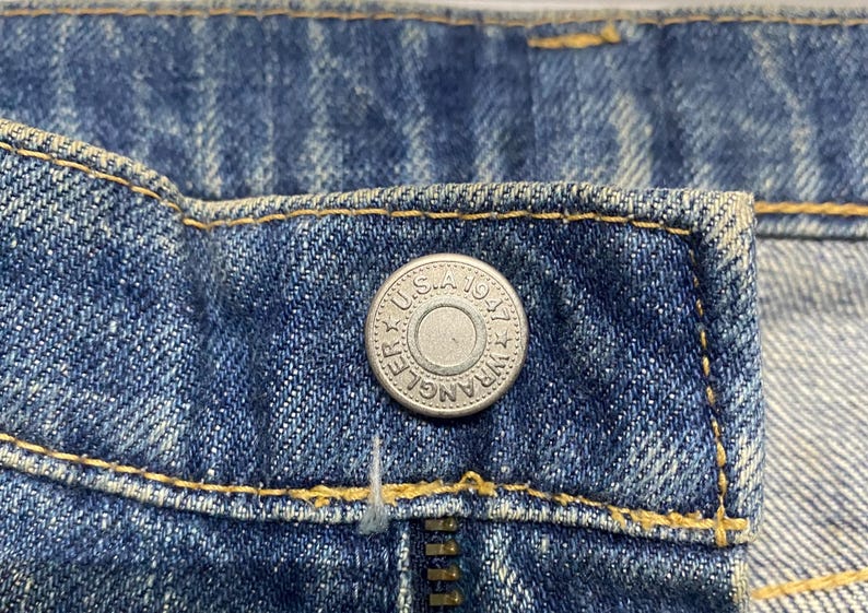 Może przedstawiać: Zbliżenie niebieskich dżins&oacute;w z srebrnym guzikiem i zamkiem błyskawicznym. Na guziku widnieje napis "Wrangler U.S.A. 1947". Dżinsy mają ż&oacute;łte szwy.
