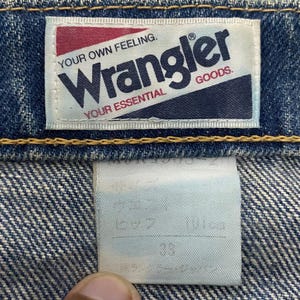 Może przedstawiać: Zbliżenie niebieskich dżins&oacute;w z etykietą Wrangler. Etykieta jest biała z czerwonym i granatowym tekstem, kt&oacute;ry brzmi "Wrangler GOODS. YOUR OWN FEELING. YOUR ESSENTIAL."