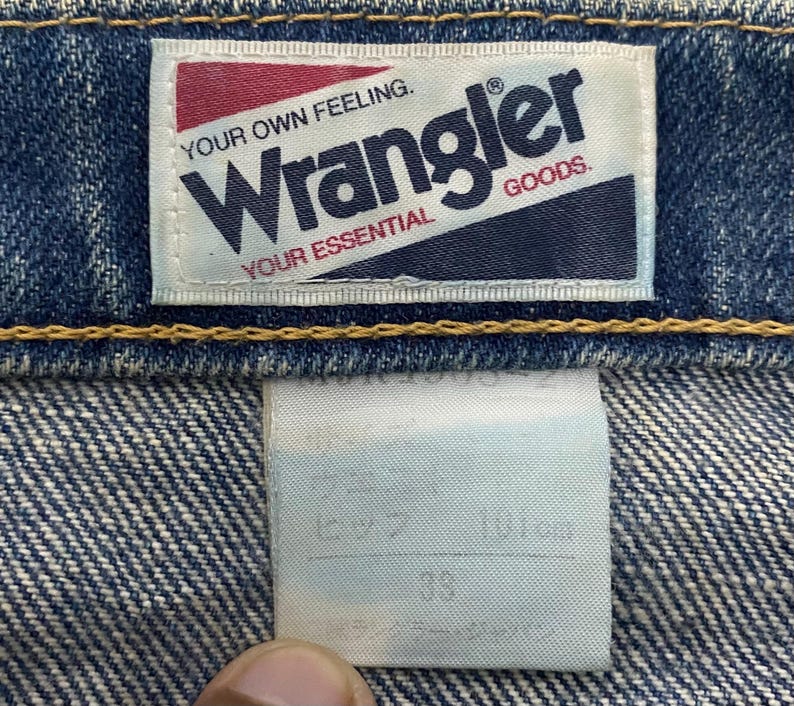 Może przedstawiać: Zbliżenie niebieskich dżins&oacute;w z etykietą Wrangler. Etykieta jest biała z czerwonym i granatowym tekstem, kt&oacute;ry brzmi "Wrangler GOODS. YOUR OWN FEELING. YOUR ESSENTIAL."