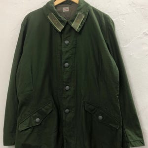 Puede incluir: Chaqueta verde oscuro de estilo militar con cierre de botones, dos bolsillos delanteros y cuello con ribete decorativo. La chaqueta está colgada de una percha de madera.