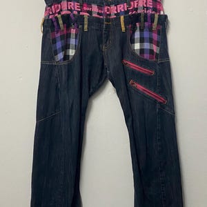 May include: Dark blue denim jeans featuring a pink and black plaid waistband with the text RIDERE sorridere. The pants include plaid pocket accents, multiple belt loops in varying colors, and two diagonal zippered pockets on the left thigh area.