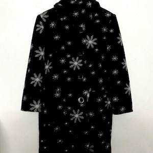 May include: Black coat with a floral pattern. The coat features a hood, long sleeves, and a button-up front. The floral design is in shades of gray and white, creating a contrasting effect against the black fabric. The coat is hanging on a metal hanger.