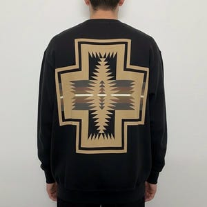 Puede incluir: Sudadera negra con un gran diseño geométrico en la espalda. El diseño tiene forma de cruz con un patrón central de zigzags y rayas horizontales en tonos marrón, beige y blanco. La sudadera es de estilo casual.
