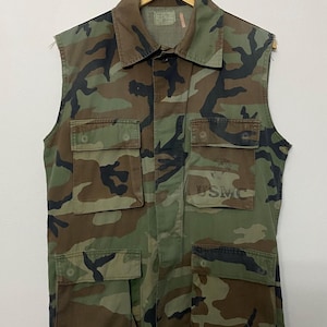 Op de afbeelding: Camouflage mouwloos vest met vier voorzakken en een kraag. Het vest heeft een groen, bruin en zwart patroon. De letters "USMC" zijn gedrukt op de rechter borstzak. Het vest hangt aan een houten hanger.
