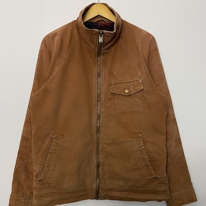 Puede incluir: Chaqueta marrón con cremallera, cuello alto y múltiples bolsillos. La chaqueta tiene una cremallera completa, dos bolsillos delanteros y un bolsillo en el pecho. Cuelga de una percha de madera sobre un fondo blanco.