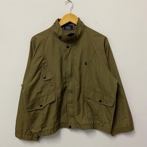 Könnte beinhalten: Olivgrüne Jacke mit Stehkragen, zwei Brusttaschen und zwei unteren Taschen. Die Jacke hat ein schwarzes Logo auf der Brust und den Text "Polo by Ralph Lauren" am Kragen.