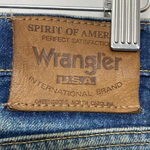 Może przedstawiać: Zbliżenie brązowej sk&oacute;rzanej metki Wrangler na niebieskich dżinsach. Na metce widnieje napis "SPIRIT OF AMERICA PERFECT SATISFACTION Wrangler USA INTERNATIONAL BRAND GREENSBORO, NORTH CAROLINA."