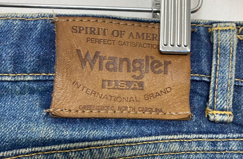 Może przedstawiać: Zbliżenie brązowej sk&oacute;rzanej metki Wrangler na niebieskich dżinsach. Na metce widnieje napis "SPIRIT OF AMERICA PERFECT SATISFACTION Wrangler USA INTERNATIONAL BRAND GREENSBORO, NORTH CAROLINA."