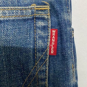 Może przedstawiać: Zbliżenie niebieskich dżins&oacute;w z ż&oacute;łtymi szwami. Czerwona prostokątna metka z białym napisem "WRANGLER" jest wszyta na tylnej kieszeni. Dżinsy mają klasyczny, zużyty wygląd.