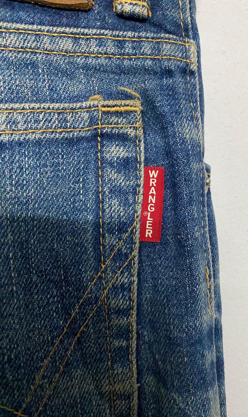 Może przedstawiać: Zbliżenie niebieskich dżins&oacute;w z ż&oacute;łtymi szwami. Czerwona prostokątna metka z białym napisem "WRANGLER" jest wszyta na tylnej kieszeni. Dżinsy mają klasyczny, zużyty wygląd.