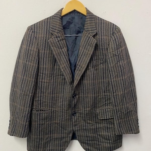May include: A brown and navy herringbone blazer with a two-button fastening and two front pockets. The jacket is suspended on a wooden hanger. The blazer has a dark blue lining. The background is a white wall.