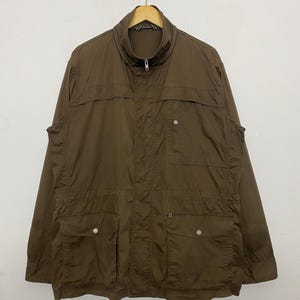 May include: A brown jacket with a high collar, a zip fastening, and several pockets. The jacket is suspended on a wooden hanger against a plain white backdrop. The jacket has two chest pockets and two lower pockets.