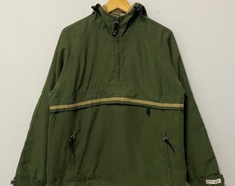 May include: Olive green anorak jacket with a tan horizontal stripe across the chest. The jacket has a hood, a half-zip closure, and two front pockets. The cuffs have a small logo patch. The jacket is hanging on a wooden hanger.