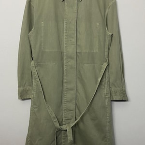 May include: A long-sleeved, sage green trench coat featuring a classic collar, a concealed button-front placket, and a matching fabric tie belt at the waist. The coat has a structured silhouette with two large rectangular chest pockets and long sleeves.
