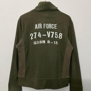 May include: Olive green zip-up jacket with a high collar. The back features white block lettering that reads "AIR FORCE" above "274-V758" and "GS8N 8-15". The jacket has brown panels on the sides and is hanging from a metal hanger.