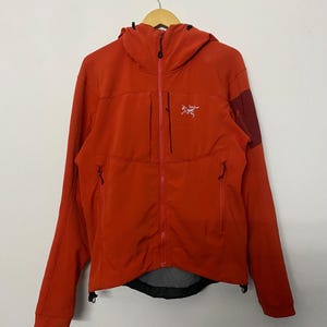 Puede incluir: Una chaqueta con capucha de color naranja brillante que cuenta con una cremallera frontal completa, un bolsillo vertical en el pecho y dos bolsillos laterales con cremallera. La prenda muestra un logotipo blanco en el pecho izquierdo y un parche rojo oscuro en la manga izquierda.