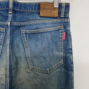 Może przedstawiać: Niebieskie dżinsy Wrangler z wyblakłym denim. Tylna kieszeń ma charakterystyczne przeszycia w kształcie "W". Widoczna jest brązowa sk&oacute;rzana naszywka z marką "Wrangler". Czerwona metka "Wrangler" znajduje się na tylnej kieszeni. Dżinsy wiszą na drewnianym wieszaku.