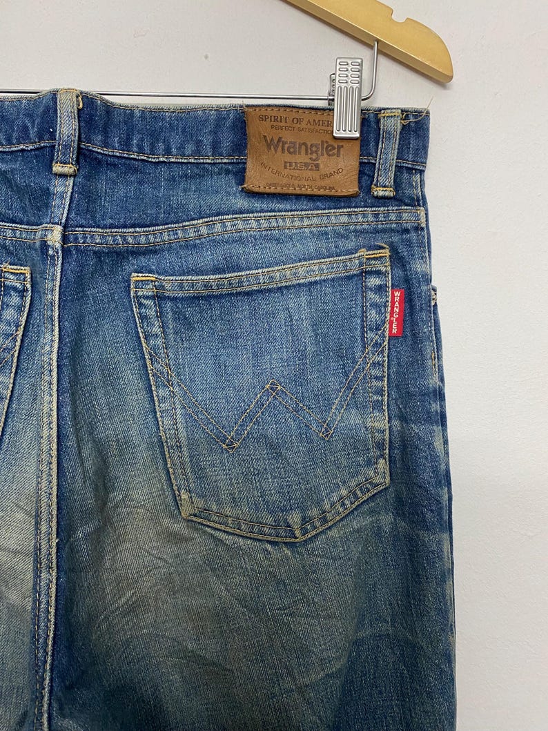 Może przedstawiać: Niebieskie dżinsy Wrangler z wyblakłym denim. Tylna kieszeń ma charakterystyczne przeszycia w kształcie "W". Widoczna jest brązowa sk&oacute;rzana naszywka z marką "Wrangler". Czerwona metka "Wrangler" znajduje się na tylnej kieszeni. Dżinsy wiszą na drewnianym wieszaku.