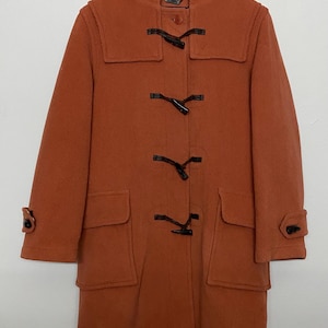 May include: A terracotta-coloured duffel coat with a hood, toggle fastenings, and two front pockets. The coat features black toggles and buttons, and is suspended on a wooden hanger. The coat is made of a thick, warm material.