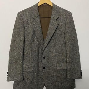 May include: A tweed blazer in shades of gray and brown hangs on a wooden hanger. The jacket features a two-button closure, notched lapels, and two front pockets. The sleeves have three buttons at the cuffs. The interior lining is a solid brown color.