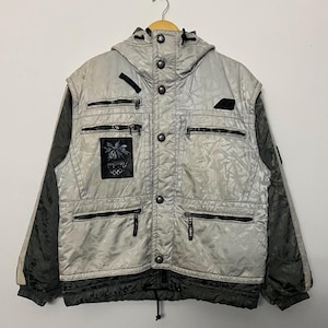 Peut inclure: Veste d'hiver argentée et grise avec capuche, plusieurs poches zippées et un écusson noir et blanc représentant un palmier et le logo des Jeux olympiques de Nagano. La veste a des accents noirs sur les manches et le bas.