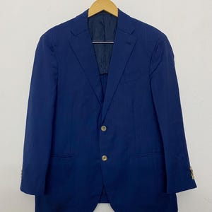 May include: A royal blue blazer featuring a subtle tonal windowpane check pattern, a notched lapel, two front buttons, and two flap pockets. The garment is lined with dark blue fabric and is displayed on a wooden hanger against a plain background.