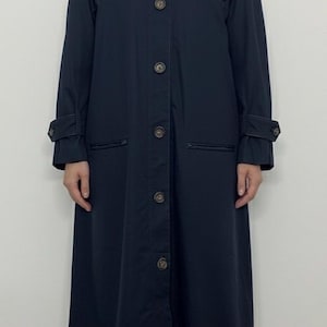 Puede incluir: Un abrigo tipo trench de color azul marino con cuello redondo, cierre de botones y mangas largas. El abrigo tiene dos bolsillos delanteros y puños abotonados. Es un estilo largo y clásico.
