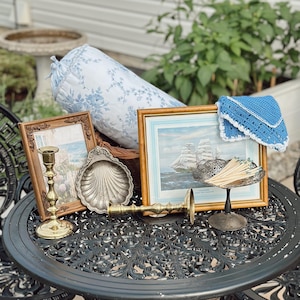 May include: A black metal bistro table with two matching chairs, featuring a blue and white floral bolster pillow, two framed pictures, a silver shell dish, a blue crocheted cloth, two brass candlesticks, and a silver bowl filled with wooden matches.
