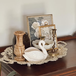 May include: A decorative silver tray holds a collection of vintage items. A white ceramic swan, two framed floral prints, a marble-like vase, a dark brown jar, a white bowl, and two brass candlesticks are arranged on the tray.